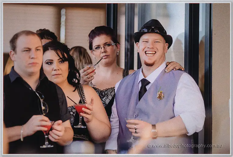Groomsmen and bridesmaids laugh over cocktails in black lace and trilby hats