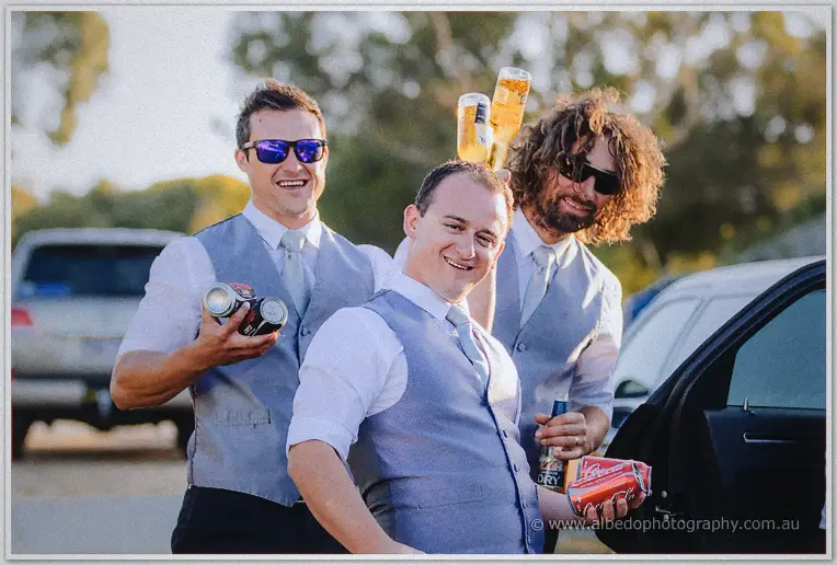 Groomsmen in grey vests and sunglasses laugh with raised beers beside cars in golden coastal light