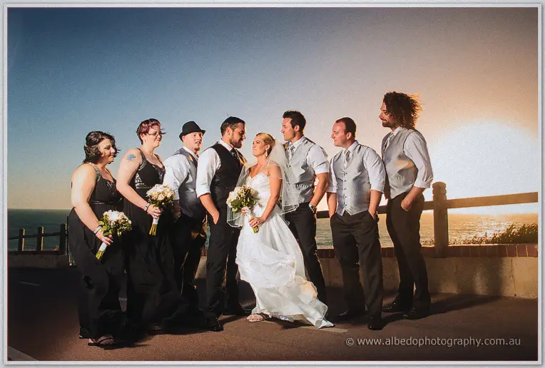 Bridal party in black and grey lines up along the sunset rail above the ocean, bouquets in hand
