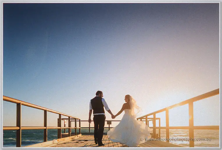 Teigan and Troy walk hand in hand down a wooden jetty, her veil streaming in the golden sea breeze