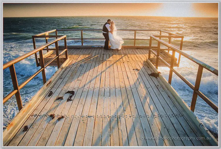 Wide view of the couple embracing at the jetty's end as waves break and long shadows stretch at sunset