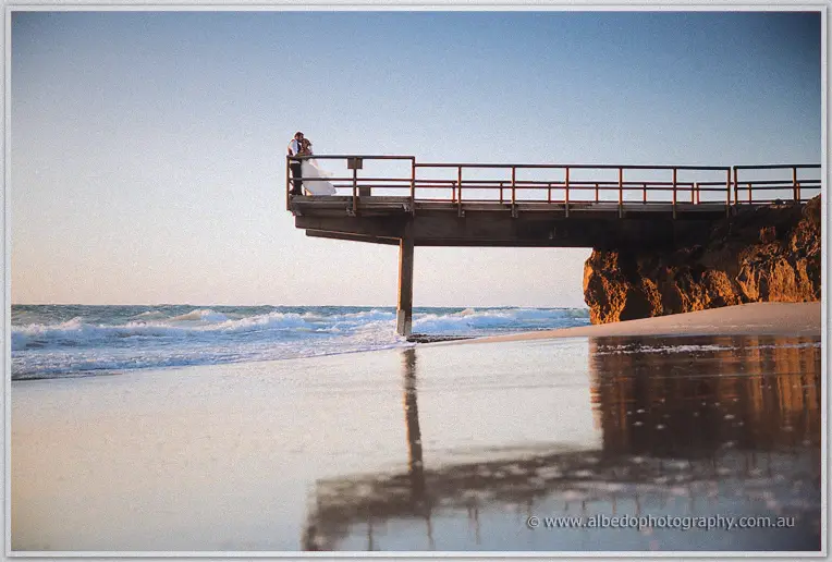 Wide beach scene with the pair far out on a jetty above wet reflective sand and gentle surf