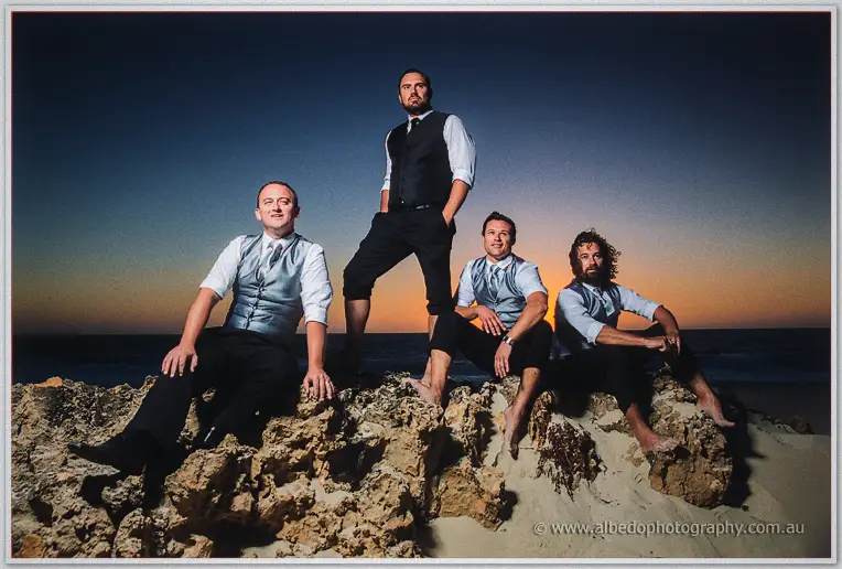 Groom and groomsmen pose barefoot on coastal rocks under a deep blue dusk sky