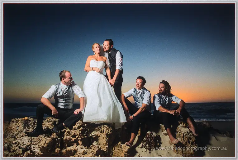 Bride and groom stand centre on the rocks flanked by laughing groomsmen at twilight