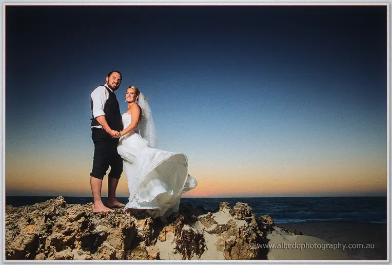 Teigan's gown billows in the wind as she and Troy hold hands on the rocks at dusk