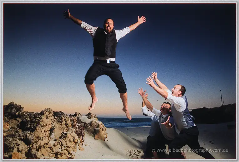 Playful groomsmen toss one leaping friend into the air on the twilight beach