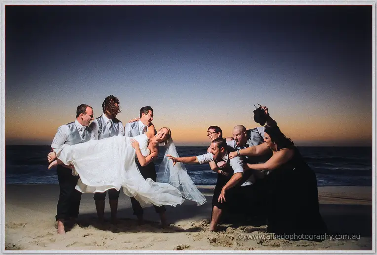 Groomsmen swing the laughing bride while bridesmaids charge in a mock tug-of-war on the dusk beach