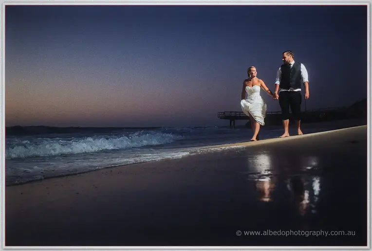 Teigan and Troy walk hand in hand along the darkening shoreline, her gown hem lifted, jetty silhouette behind