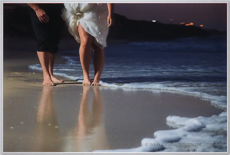 Close view of bare feet in the lapping surf edge, dusk lights glinting across the bay