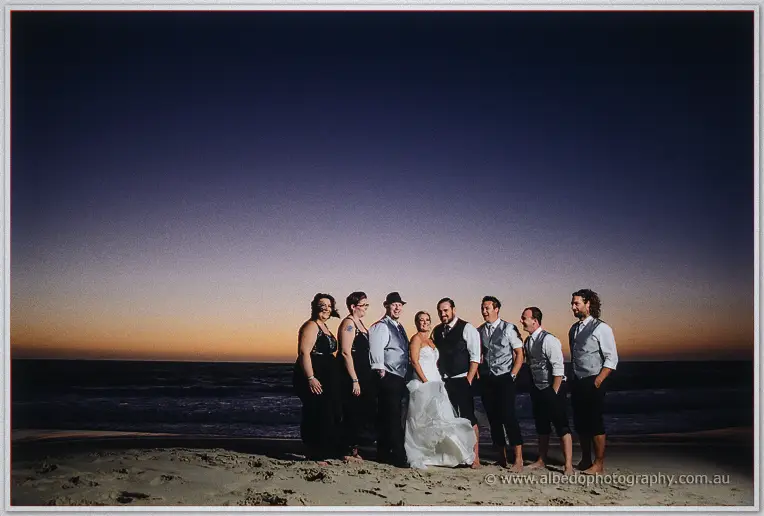 The whole bridal party lines the darkening beach beneath a wide violet afterglow sky