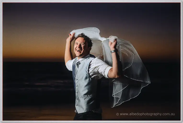 A grinning groomsman flings the bride's veil overhead like a cape on the night beach