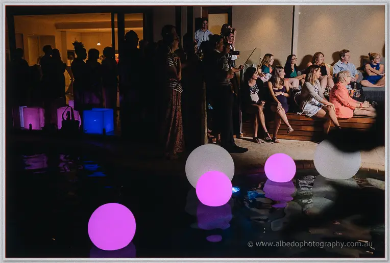 Guests crowd the pool deck as pink and white orbs glow across the water at night