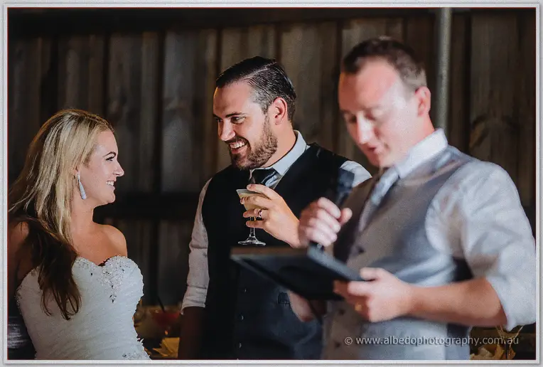 Best man raises a cocktail mid-speech while the couple trade amused looks across the barn table