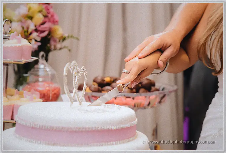 Close-up of ringed hands steering the knife into the white and pink heart-topped cake