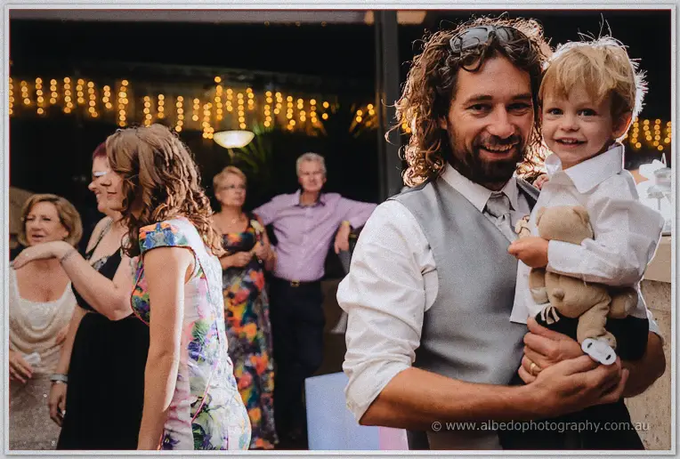 A curly-haired groomsman grins while cuddling a laughing little boy clutching a teddy bear