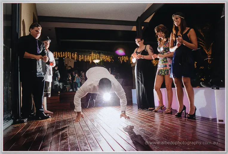 A guest drops low in a breakdance pose, hands planted, as friends cheer around the floor