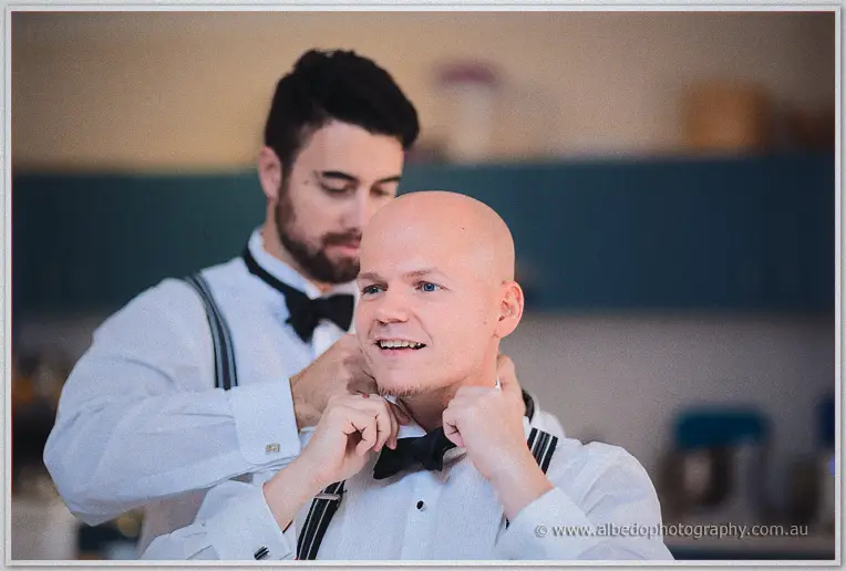 A groomsman helps Michael fasten his bow tie during morning preparations.