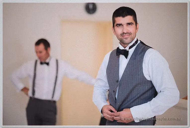 A groomsman in a striped grey vest buttons his waistcoat as a colleague adjusts his braces behind.