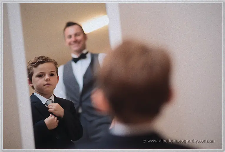 The young page boy checks his tie in the mirror while a groomsman smiles behind him.