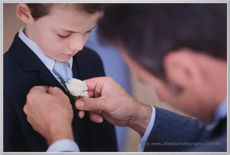A groomsman pins a white rose boutonniere onto the page boy's dark suit jacket.