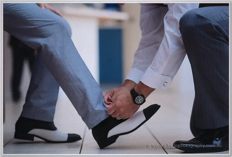 A groomsman crouches to help Michael into his two-tone spectator shoes.
