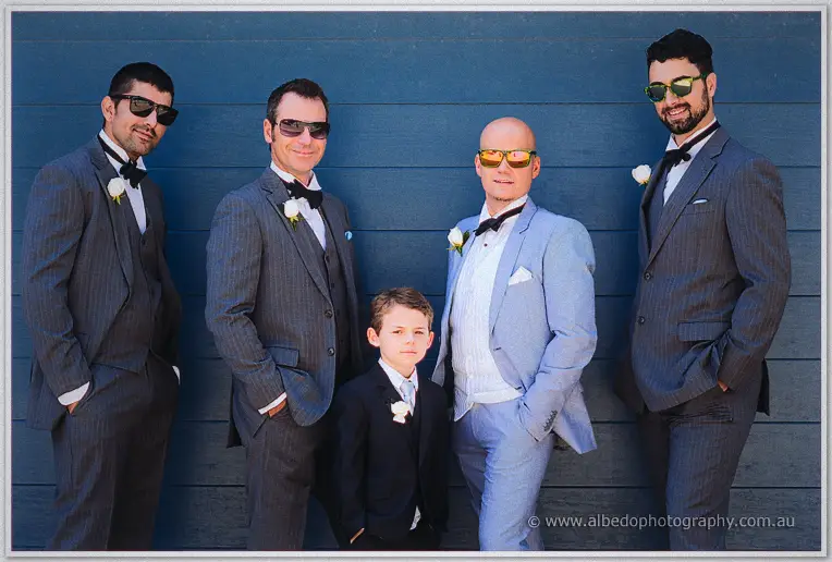 Michael, groomsmen and page boy pose in sunglasses against a deep blue weatherboard wall.