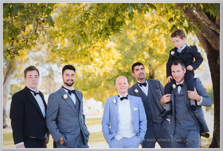 The page boy rides a groomsman's shoulders among the bridal party under golden leaves.