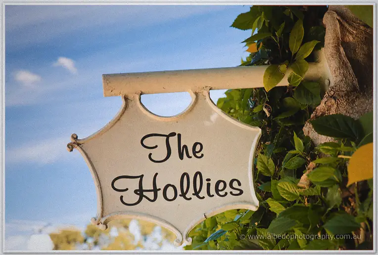 A cream fretwork sign reading The Hollies hangs among green leaves against the sky.