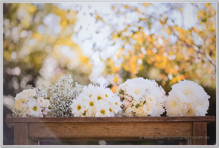 Cream wedding bouquets of roses, daisies and baby's breath on a rustic timber table under sunlit trees
