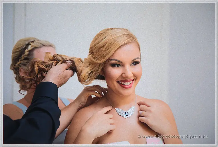 Beaming bride Jasmine laughing as stylists finish her curls and clasp the crystal wing necklace at her neck