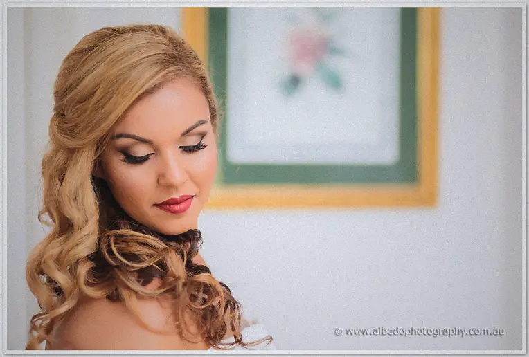 Bride Jasmine glancing down with curled golden hair, red lipstick and soft window light in a bright prep room