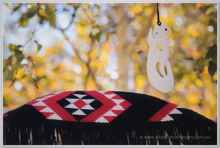 Carved bone hei matau pendant above a woven red, black and white korowai cloak, autumn leaves glowing behind