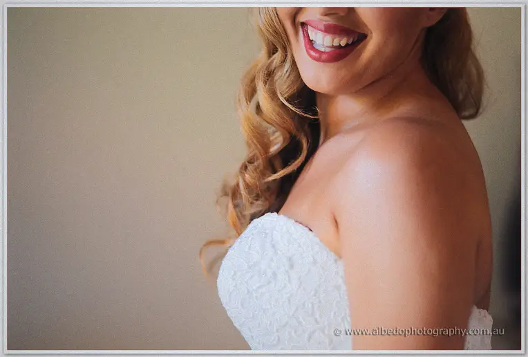 Close crop of the smiling bride's red lips and bare shoulder above her strapless lace bodice