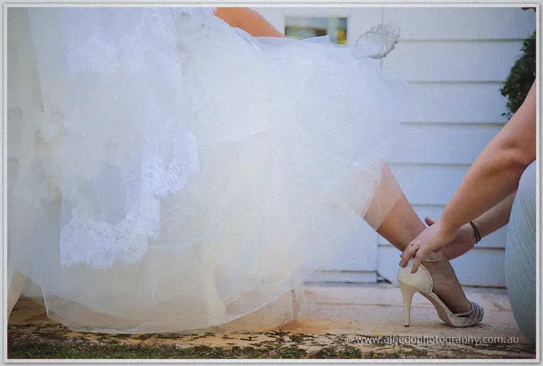 Bridesmaid kneeling to slip a glittering heel onto the bride's foot beneath billowing white tulle
