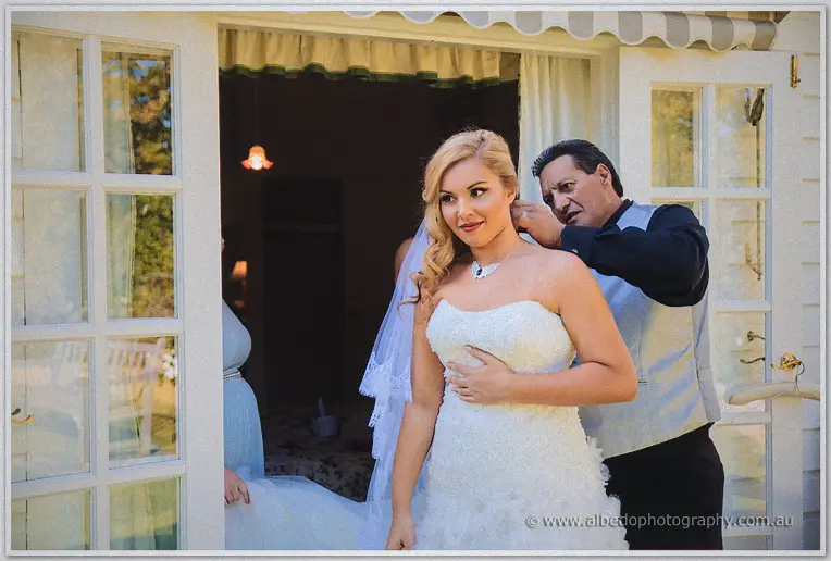 Veiled bride Jasmine pausing at French doors as her father fastens her necklace before the ceremony