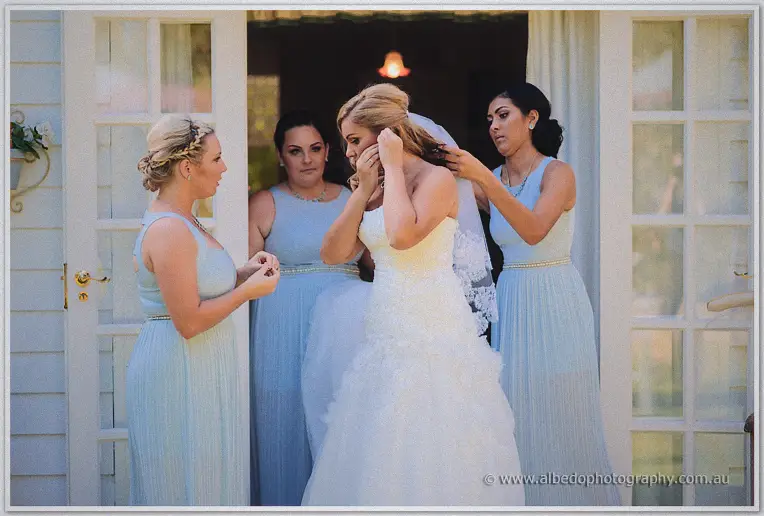 Bridesmaids in powder-blue gowns crowding around Jasmine to pin her veil at the doorway of a white cottage