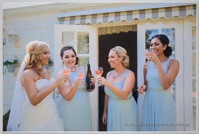Bride Jasmine and her bridesmaids in pale blue toasting with rose sparkling wine by the cottage doors