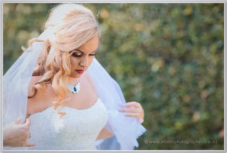 Bride Jasmine arranging her flowing veil while sunlight rims her curls against soft green foliage