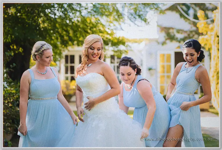 Bridesmaids in sky blue gathered around Jasmine laughing as they arrange her gown on the Sunalford lawn