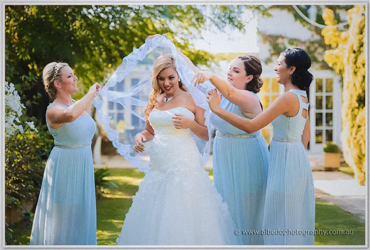 Bridesmaids lifting Jasmine's lace-edged veil into the breeze as the group giggles beneath golden garden trees