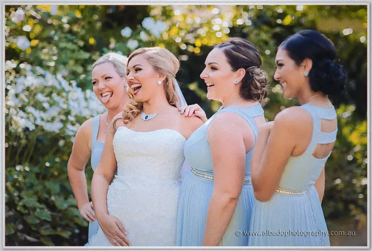 Playful candid of the bridal party in powder blue pulling faces behind a laughing Jasmine amid garden blooms