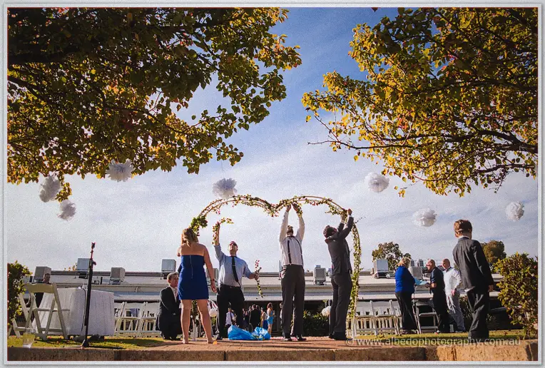 Men in braces raising a greenery wedding arbour under autumn trees while guests gather for the ceremony