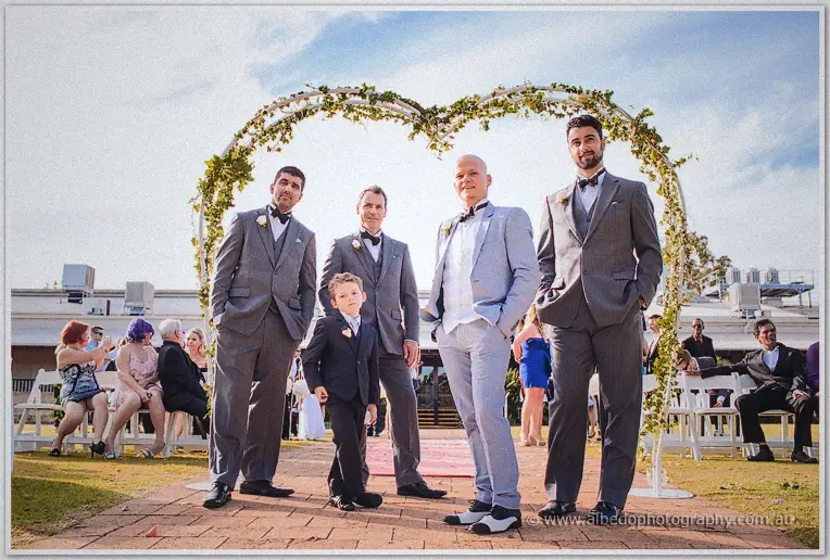 Groom Michael in a pale grey suit flanked by groomsmen and a young page boy beneath the heart-shaped ivy arch