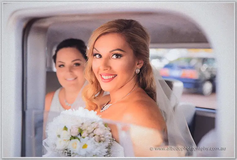Bride Jasmine beaming from the limousine window clutching her white bouquet, bridesmaid smiling behind