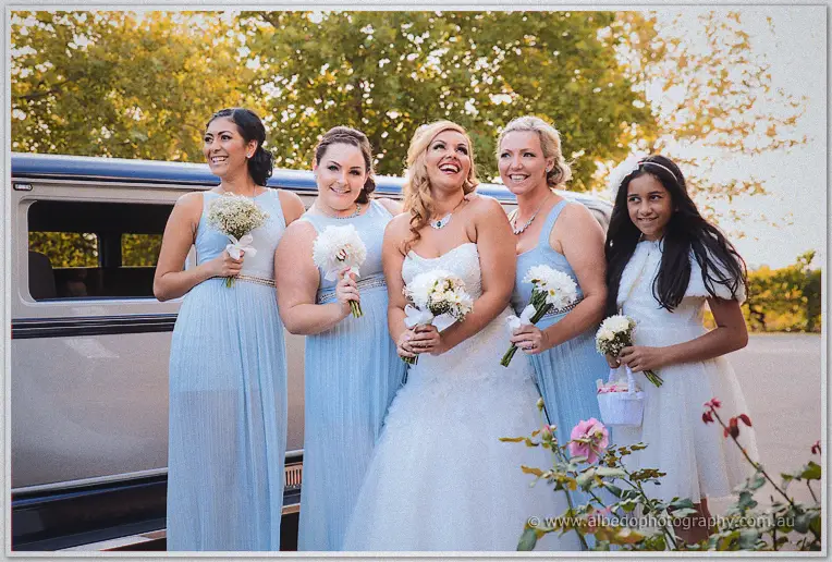 Jasmine laughing with her bridesmaids and flower girl holding baby's breath posies in front of the limousine