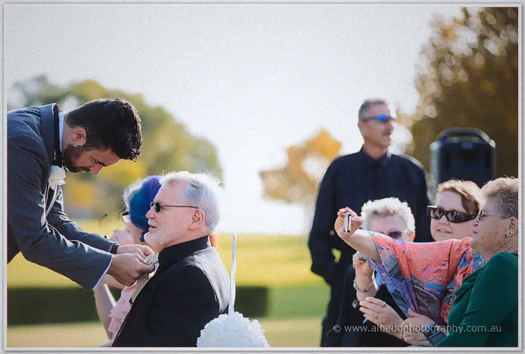 Groomsman pinning a boutonniere on a seated grey-bearded guest as family chat in the warm afternoon light