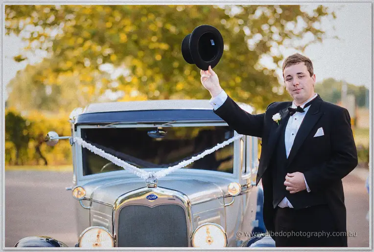 Chauffeur in black tie raising his top hat beside the ribbon-trimmed vintage bridal car among golden trees