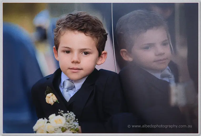 Pensive page boy in a navy suit and cream rose boutonniere holding white posies, reflected in the car window