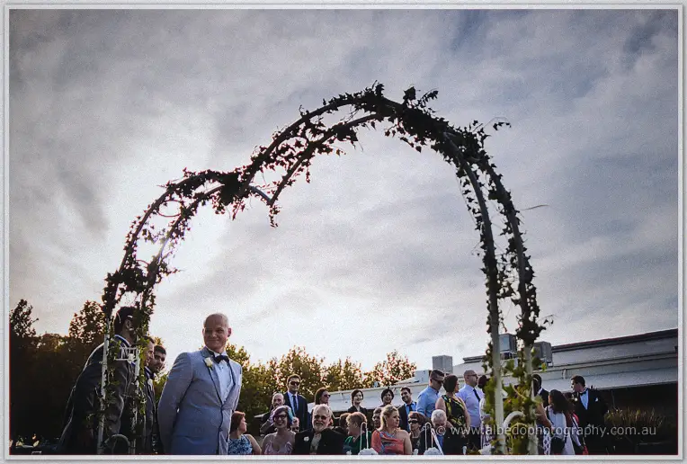 Groom Michael grinning under the ivy heart arch against a moody dusk sky as guests gather