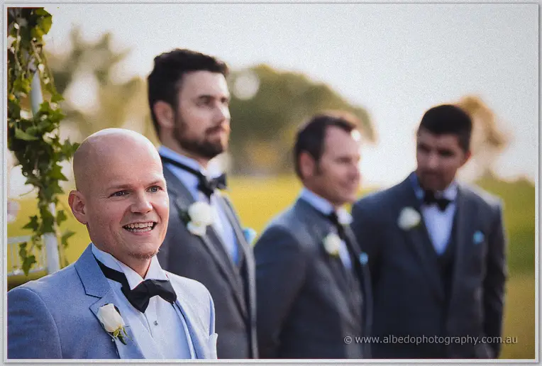 Groom Michael beaming as he waits at the arbour, groomsmen in grey suits soft-focused behind him